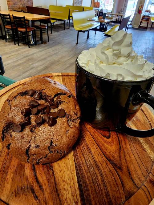 A large chocolate chip cookie next to a black mug topped with whipped cream, set on a wooden platter in a café.