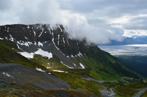 A scenic mountain landscape with snow patches, clouds, and a river winding through lush greenery below.