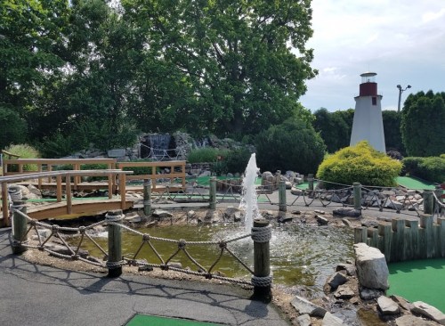 A scenic mini-golf course featuring a pond, fountain, wooden bridge, and a lighthouse in the background.