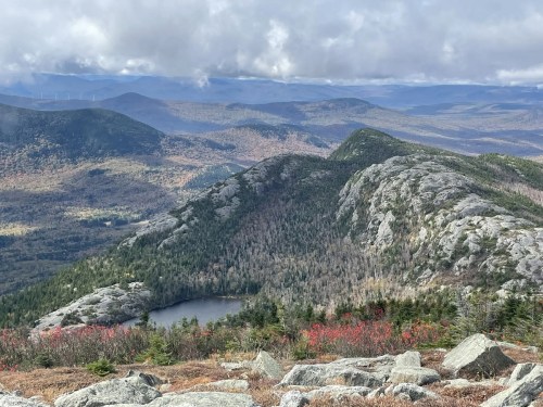 A panoramic view of mountains with autumn foliage, featuring a small lake nestled among rocky terrain.