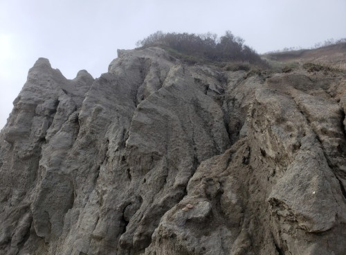 A rugged cliff face with steep, textured rock formations and sparse vegetation at the top under a cloudy sky.
