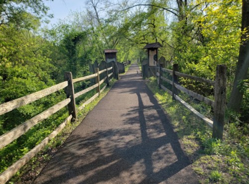 A peaceful pathway lined with wooden fences, surrounded by lush greenery and trees on a sunny day.
