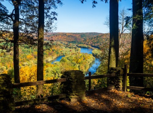 A scenic view of a river winding through colorful autumn trees, framed by tall trees and a stone railing.