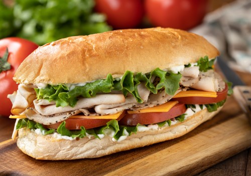 A fresh sub sandwich filled with turkey, lettuce, tomato, and cheese on a wooden cutting board, with tomatoes in the background.