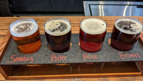 A flight of four craft beers in glasses, labeled Smoked, Porter, Buck, and Brown, on a wooden tray.