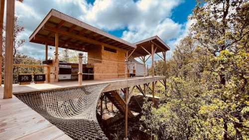 A wooden treehouse with a kitchen and hammock, surrounded by trees under a partly cloudy sky.