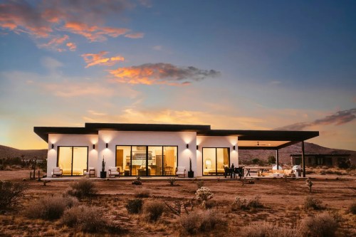 Modern desert home at sunset, featuring large windows and a spacious patio surrounded by sparse vegetation.
