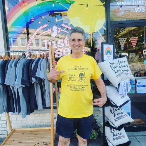 A man in a yellow shirt stands outside a store, pointing to his shirt that reads "I am a HUMAN COUPON."