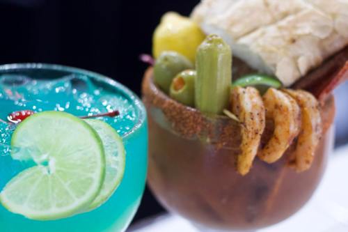 A vibrant blue cocktail with lime slices next to a savory drink garnished with shrimp, olives, and celery.