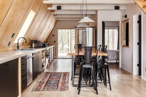 Modern kitchen and dining area with wooden accents, a large table, black chairs, and a view of nature through glass doors.