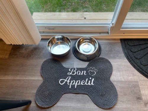 A dog mat shaped like a bone with "Bon Appetit" text, placed next to two stainless steel food bowls.