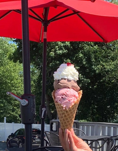 A hand holding a tall ice cream cone with three scoops: chocolate, strawberry, and topped with whipped cream and a cherry.