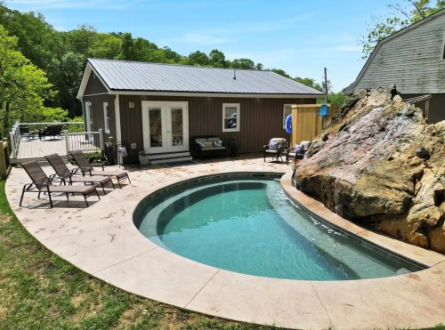A small, curved pool surrounded by lounge chairs and a rock feature, next to a cozy cabin in a green outdoor setting.
