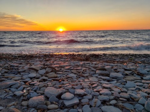 A serene beach scene at sunset, with the sun dipping below the horizon and waves gently lapping at a rocky shore.