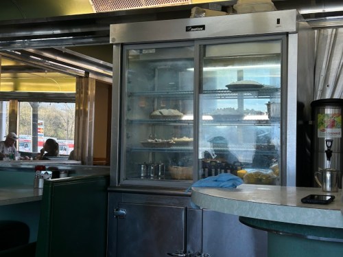 A glass display case in a diner, showcasing various baked goods and pastries, with a blurred background of patrons.