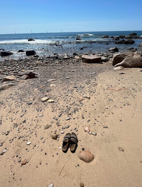 A pair of sandals on a sandy beach, with pebbles and the ocean in the background under a clear blue sky.