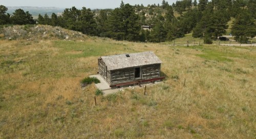 A weathered log cabin sits alone in a grassy field, surrounded by trees and hills under a clear sky.