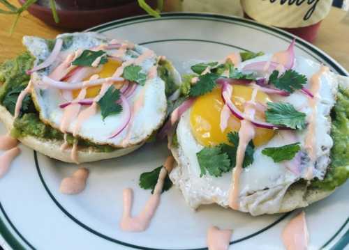 Two toasted English muffins topped with guacamole, fried eggs, red onion, cilantro, and a drizzle of sauce.