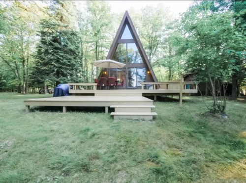 A modern A-frame cabin surrounded by trees, featuring a deck and outdoor seating area.