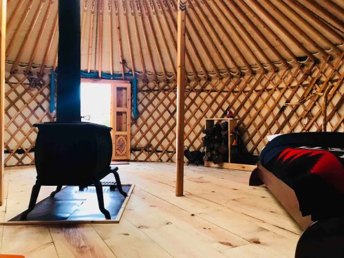 Interior of a yurt featuring a wood stove, wooden floor, and a cozy bed, with natural light coming through the door.