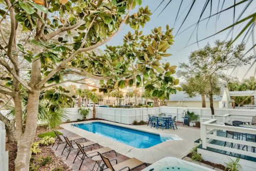 A serene backyard with a pool, surrounded by trees and lounge chairs, featuring a dining area nearby.