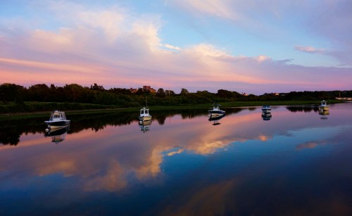 Boats anchored on a calm river at sunset, reflecting colorful clouds and greenery in the water.