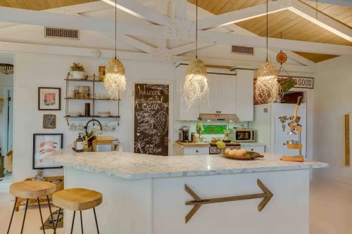 Bright kitchen with a marble island, wooden stools, pendant lights, and a chalkboard wall, featuring modern decor and greenery.