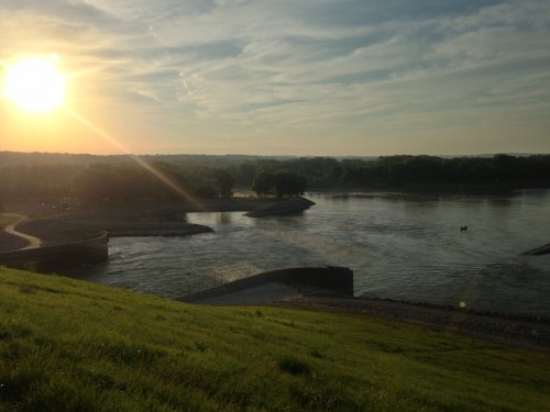 Sunset over a river, with green hills in the foreground and trees lining the water's edge.