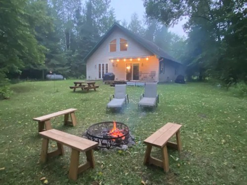 A cozy outdoor scene with a fire pit surrounded by benches, leading to a house in a wooded area on a foggy evening.
