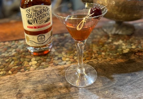 A cocktail in a martini glass with a cherry garnish, next to a bottle of Sudden Wisdom rye whiskey on a wooden surface.