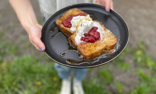 A person holds a black plate with two slices of French toast topped with whipped cream and raspberry sauce, drizzled with syrup.
