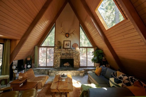 Cozy A-frame cabin interior with a stone fireplace, large windows, and wooden accents, surrounded by nature.