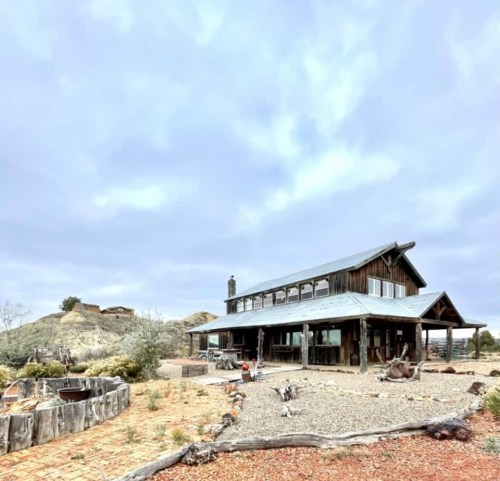 A rustic wooden house with a metal roof, surrounded by gravel and desert landscape under a cloudy sky.