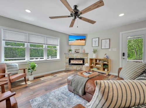 Cozy living room with a stone fireplace, TV, wooden furniture, and large windows overlooking greenery.
