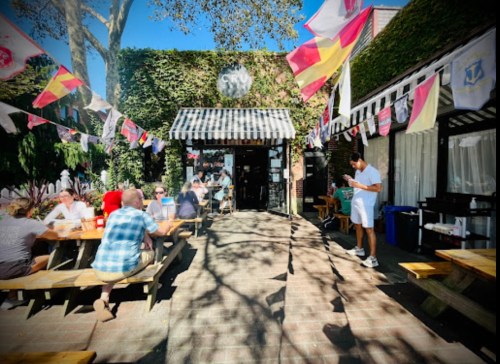 A lively outdoor dining area with people at picnic tables, colorful flags, and a server attending to guests.