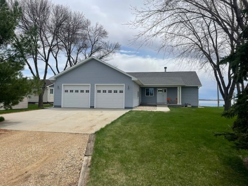 A gray house with a double garage, surrounded by green grass and trees, near a body of water under a cloudy sky.