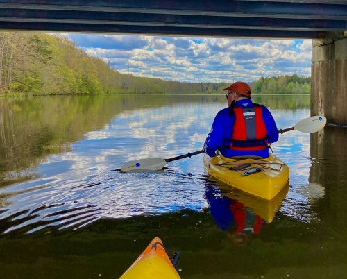 A person in a yellow kayak paddles under a bridge, surrounded by calm water and trees reflecting in the sky.