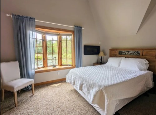 Cozy bedroom with a large window, a bed with white linens, a wooden nightstand, and a chair by the window.