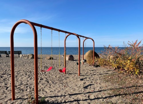 A playground swing set on a sandy beach, with the ocean and blue sky in the background.