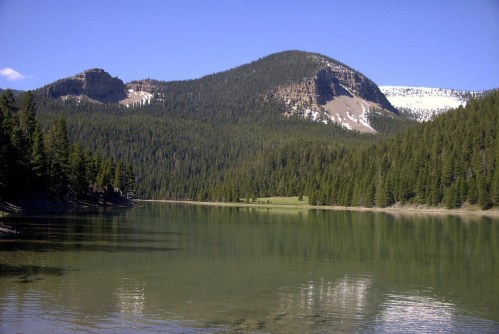 A serene lake surrounded by lush green trees and mountains under a clear blue sky. Snow-capped peaks in the distance.