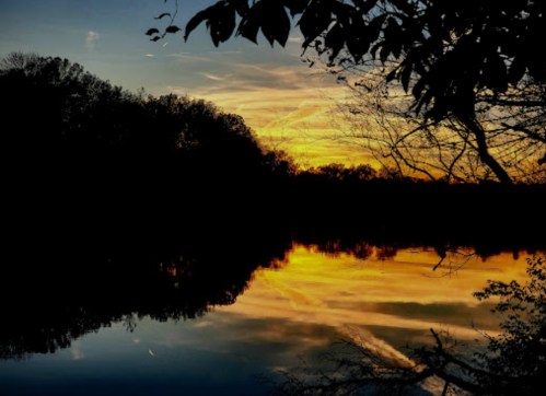 A serene sunset over a calm lake, with vibrant colors reflecting on the water and silhouetted trees in the foreground.