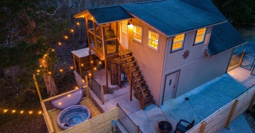 Aerial view of a cozy cabin with a hot tub, string lights, and a staircase leading to a second floor.