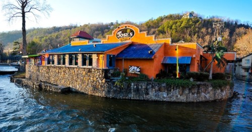 Colorful restaurant on a stone foundation by the water, surrounded by trees and hills in the background.