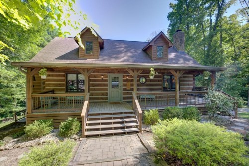 A charming log cabin surrounded by trees, featuring a spacious porch and stone pathway.