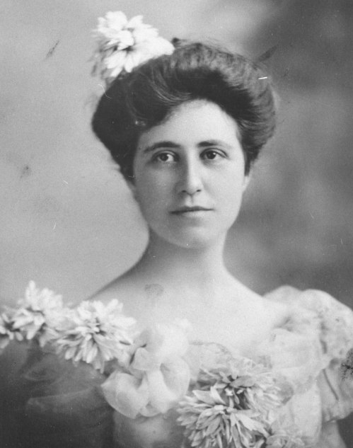 A vintage portrait of a woman with a floral hairpiece, wearing a dress adorned with large flowers, gazing directly at the camera.