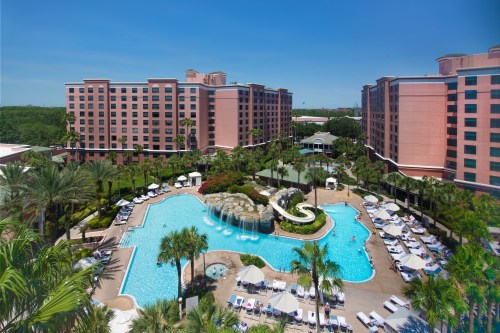 Aerial view of a resort pool surrounded by palm trees and lounge chairs, with pink buildings in the background.