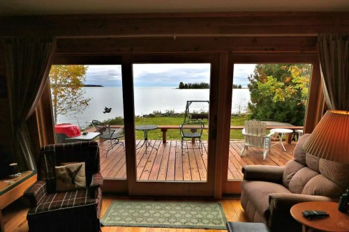 Cozy interior view of a lakefront cabin with large windows, showcasing a deck and serene water landscape outside.