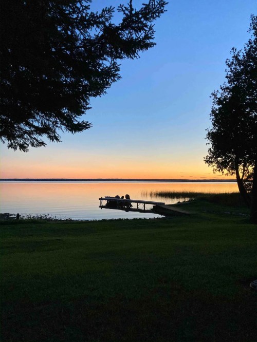 Silhouette of two people sitting on a dock by a calm lake at sunset, surrounded by trees and tall grass.