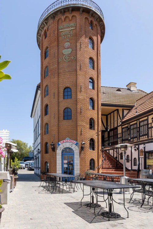 A brick tower building with large windows, surrounded by outdoor seating and a traditional-style structure nearby.