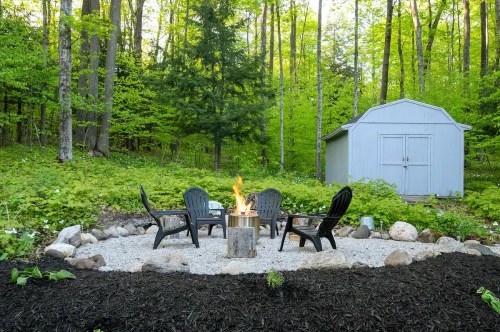 A cozy fire pit surrounded by chairs in a lush green forest, with a shed in the background.